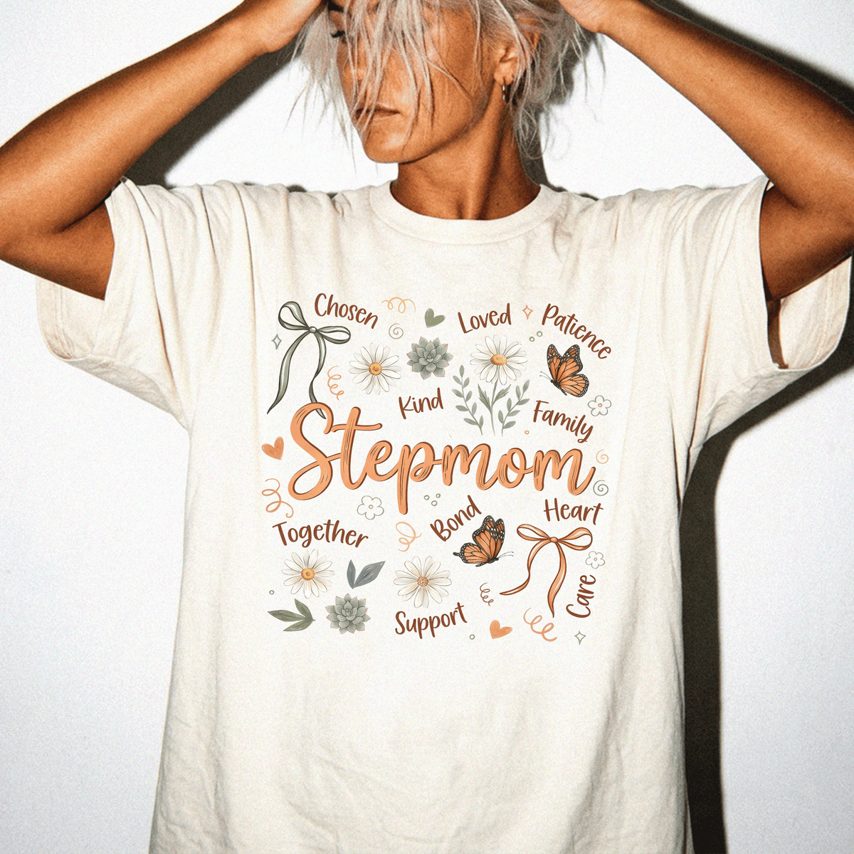 A woman wearing a white t-shirt with a graphic design featuring the word "Stepmom" surrounded by various words and illustrations such as " Chosen", "Loved", "Patience", "Kind", "Family", "Together", "Support", "Love", "CARE", and "Career".