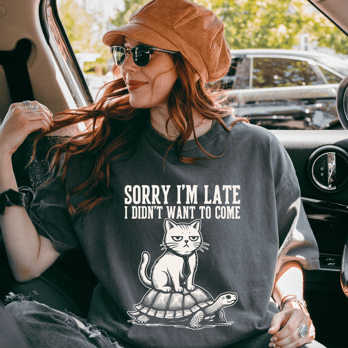 A woman wearing a hat and sunglasses is sitting in a car, looking out the window. She is wearing a gray t-shirt with a graphic design of a cat on a turtle.