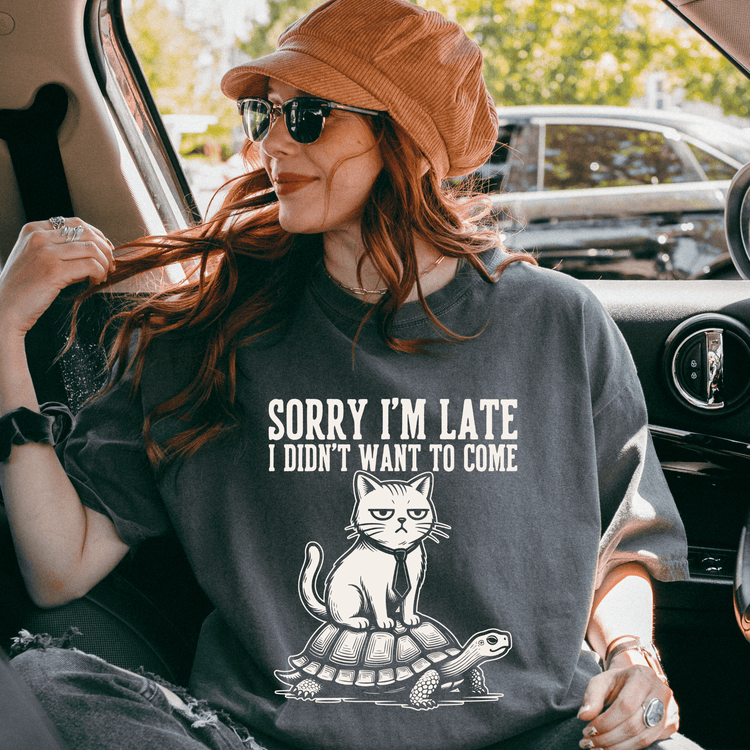 A woman wearing a hat and sunglasses is sitting in a car, looking out the window. She is wearing a gray t-shirt with a graphic design of a cat on a turtle.