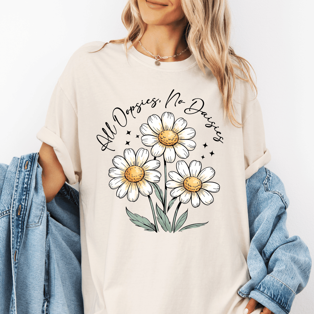 A woman wearing a white t-shirt with a graphic design of three white daisies and green leaves, along with the text "All Dopsies No Daisies" printed on it.