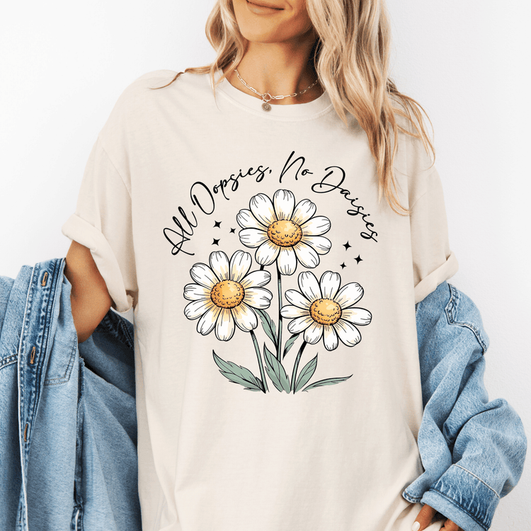 A woman wearing a white t-shirt with a graphic design of three white daisies and green leaves, along with the text "All Dopsies No Daisies" printed on it.