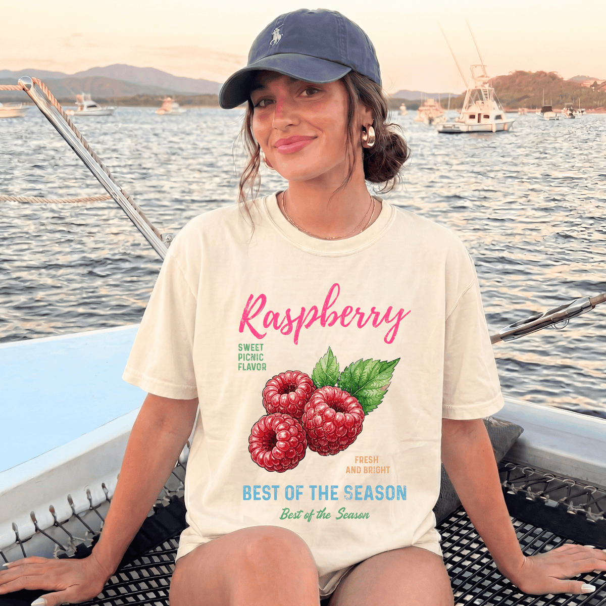 A woman wearing a white t-shirt with a raspberry graphic and text, sitting on a boat with a scenic background of water and boats.