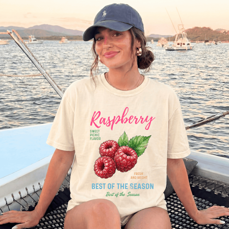 A woman wearing a white t-shirt with a raspberry graphic and text, sitting on a boat with a scenic background of water and boats.