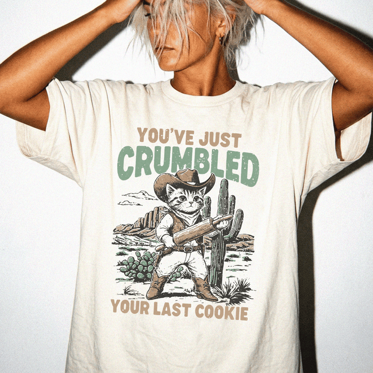 A person wearing a white t-shirt with a graphic design depicting a cat holding a rolling pin and the text "You've just Crumbled Your Last Cookie".