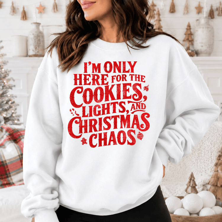 A woman wearing a white sweatshirt with a red text design that says "I'm only here for the cookies, lights, and Christmas chaos".