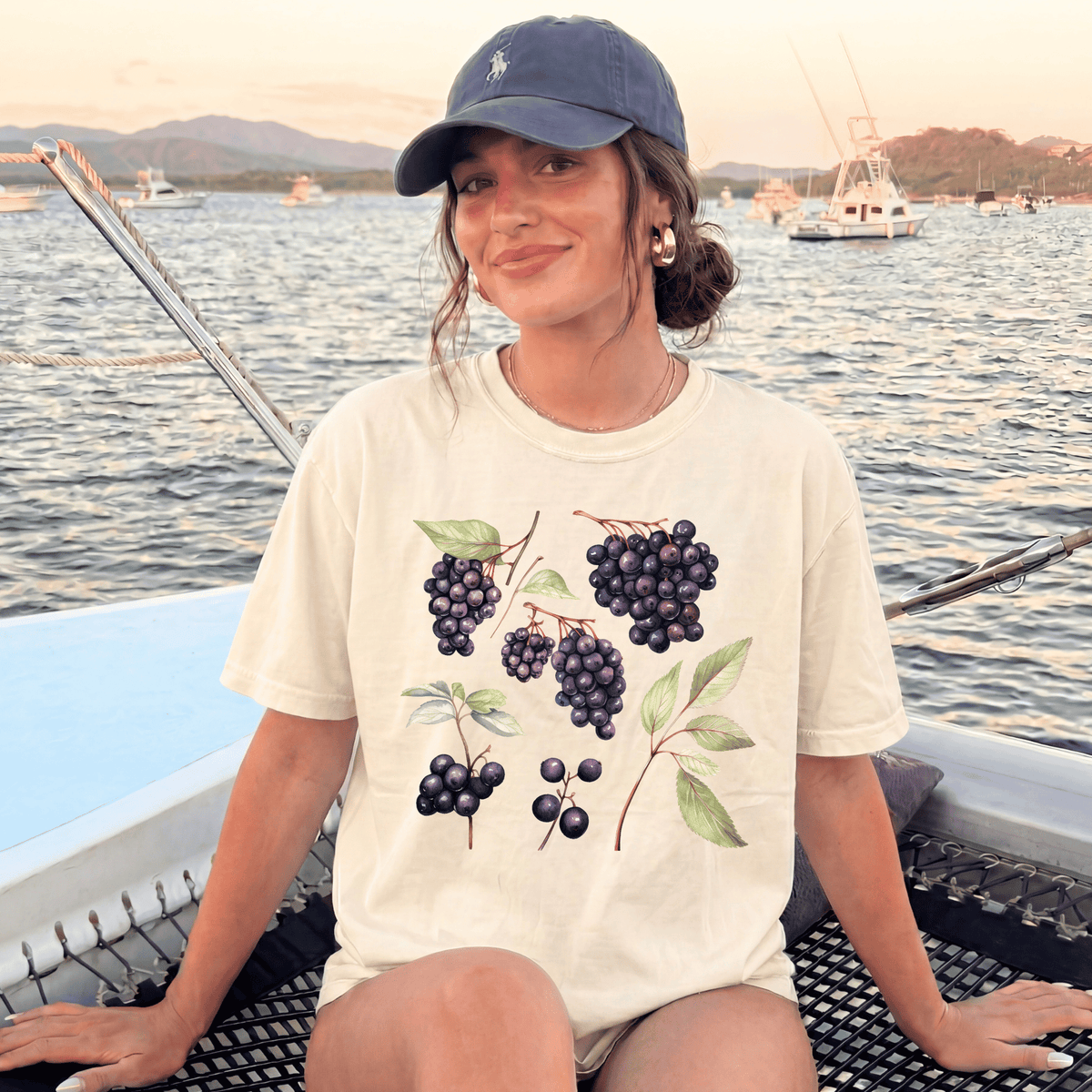 A woman wearing a white t-shirt with a graphic of grapes and leaves is sitting on a boat, smiling at the camera.