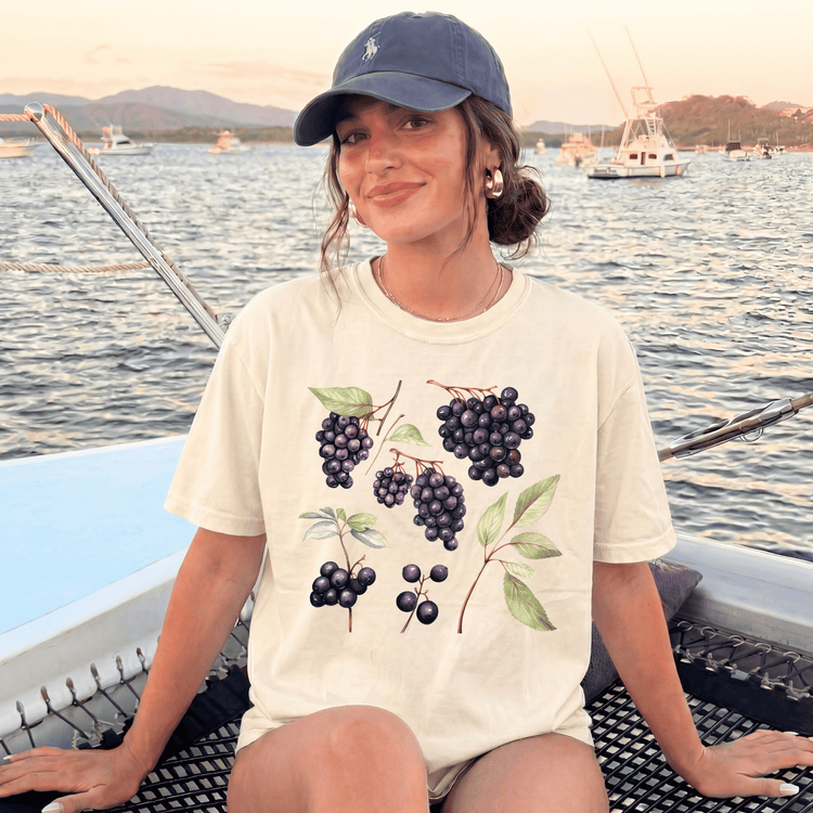 A woman wearing a white t-shirt with a graphic of grapes and leaves is sitting on a boat, smiling at the camera.