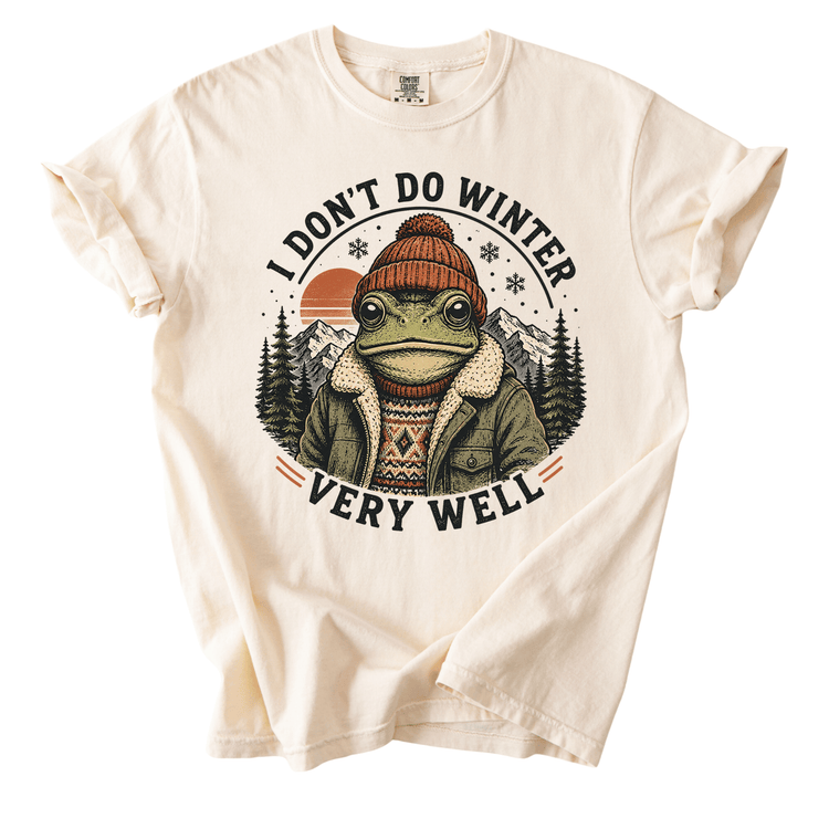A white t-shirt with a graphic design featuring a frog wearing winter clothing and a hat, surrounded by snowflakes and mountains.