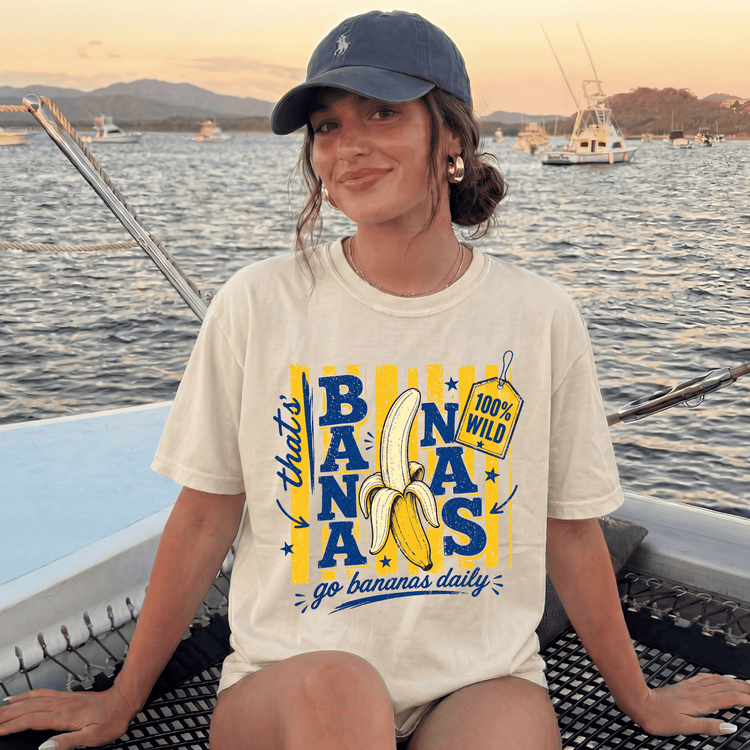A woman wearing a white t-shirt with a graphic of a banana and the text "That's bananas go bananas daily" is sitting on a boat in the water.