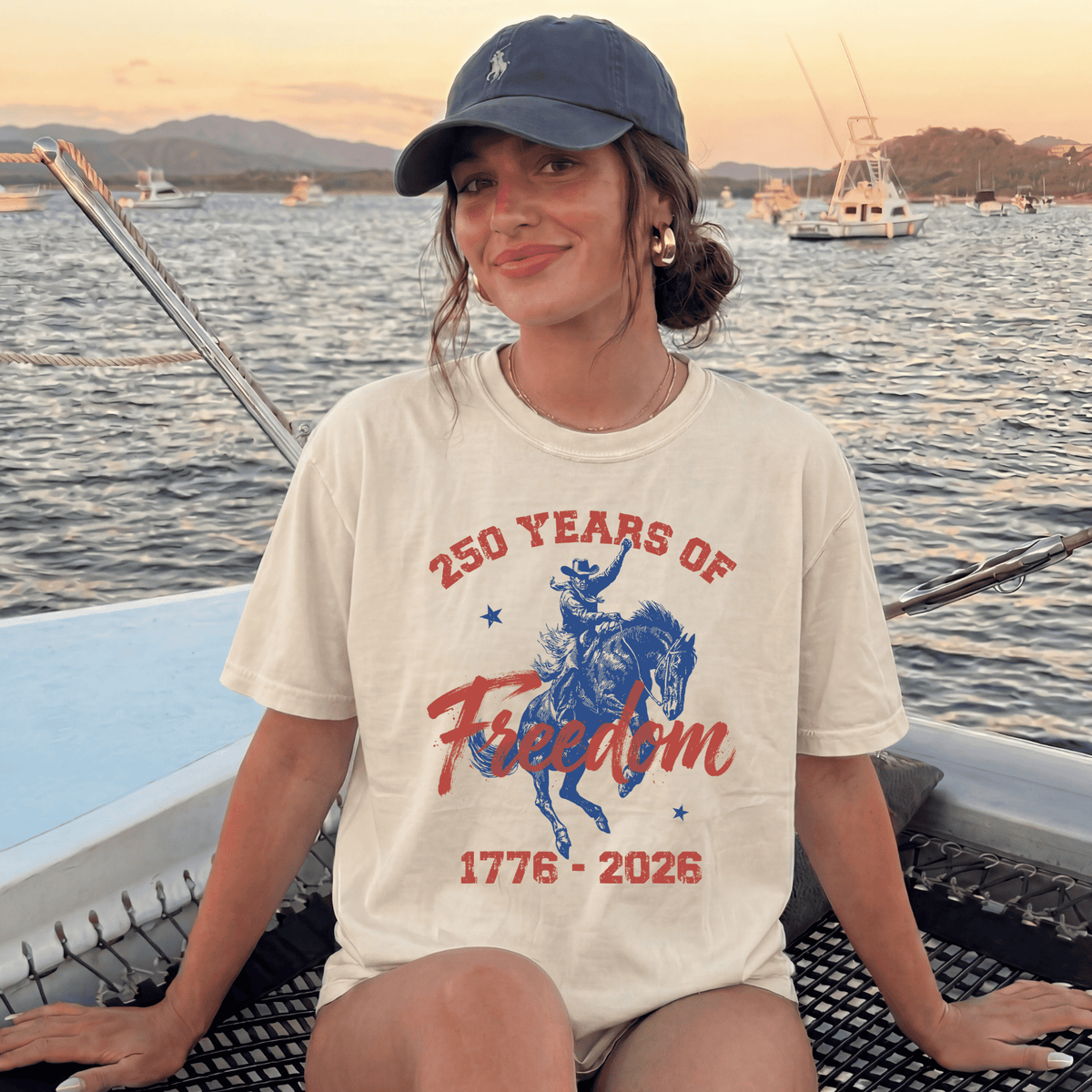 A woman wearing a cap and a t-shirt with a graphic of a cowboy on a horse, sitting on a boat in the water.
