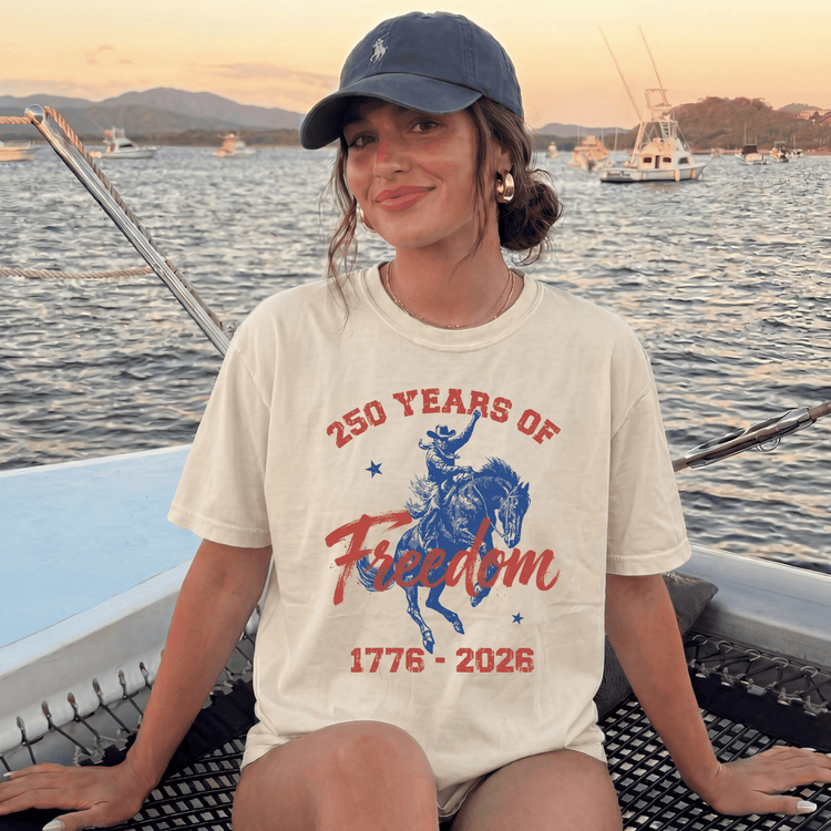 A woman wearing a cap and a t-shirt with a graphic of a cowboy on a horse, sitting on a boat in the water.