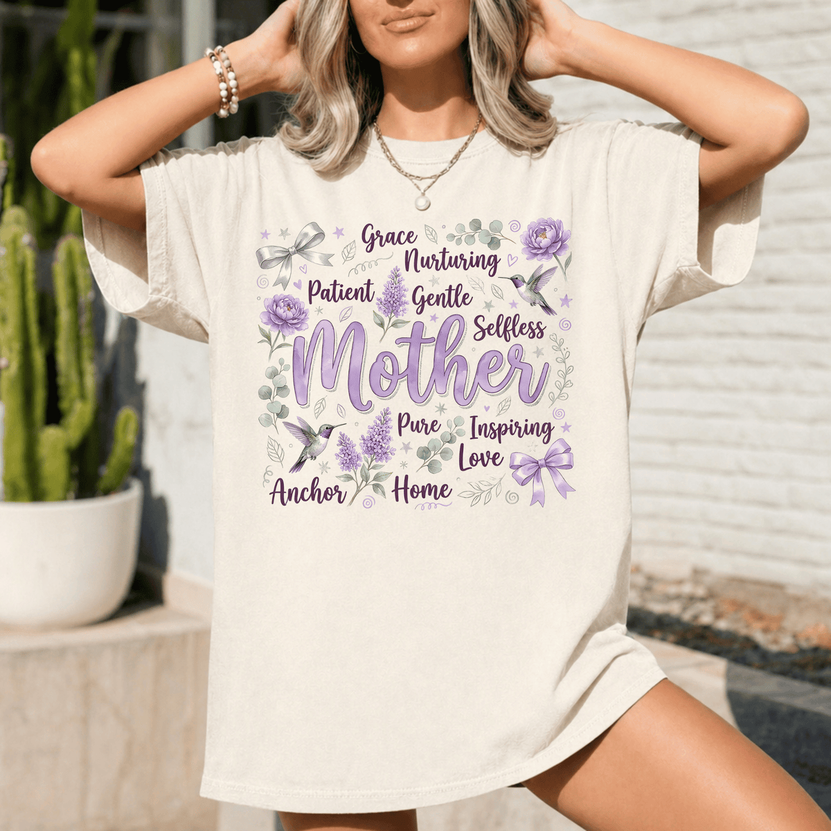 A woman wearing a white t-shirt with a graphic design featuring the words "Grace, Nurturing, Patient, Gentle, Selfless, Pure, Inspiring, Love" surrounded by purple flowers and birds.