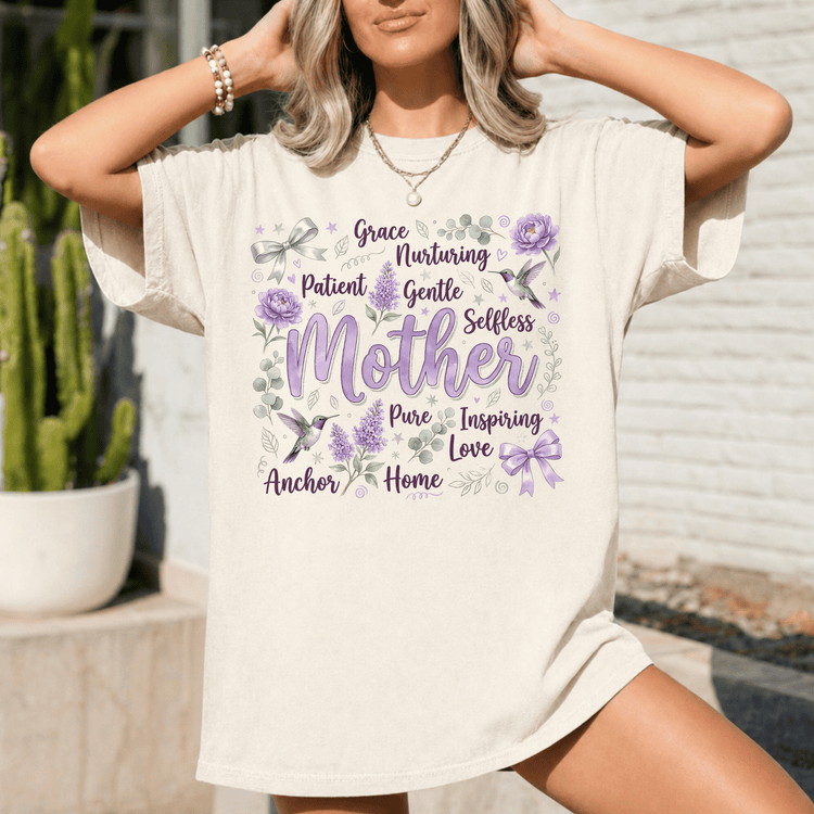 A woman wearing a white t-shirt with a graphic design featuring the words "Grace, Nurturing, Patient, Gentle, Selfless, Pure, Inspiring, Love" surrounded by purple flowers and birds.