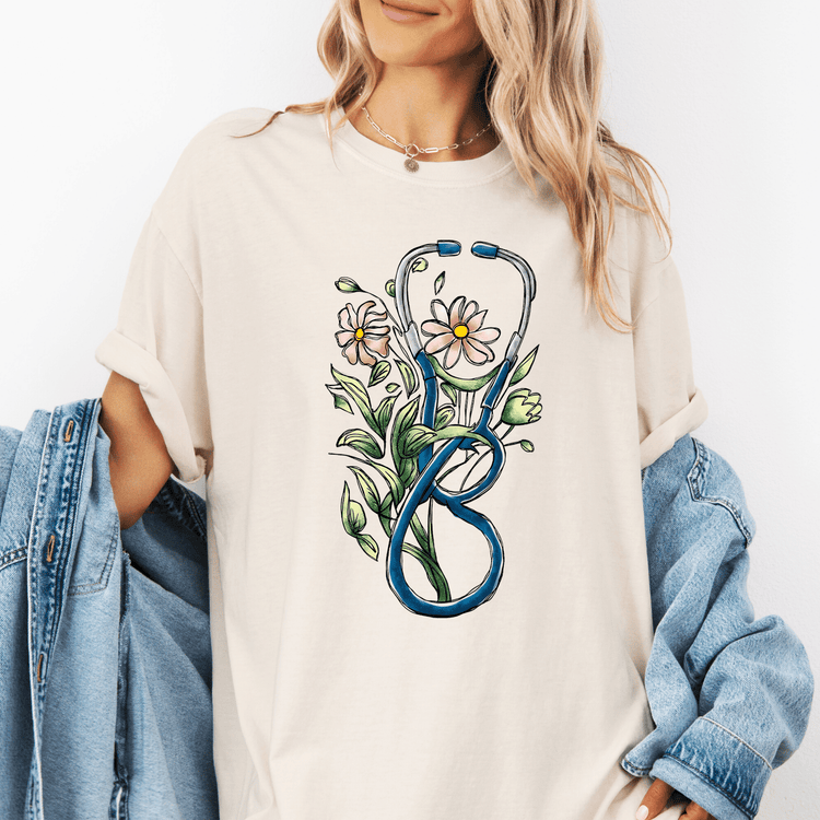 A woman wearing a white t-shirt with a graphic design of a stethoscope surrounded by flowers and leaves.