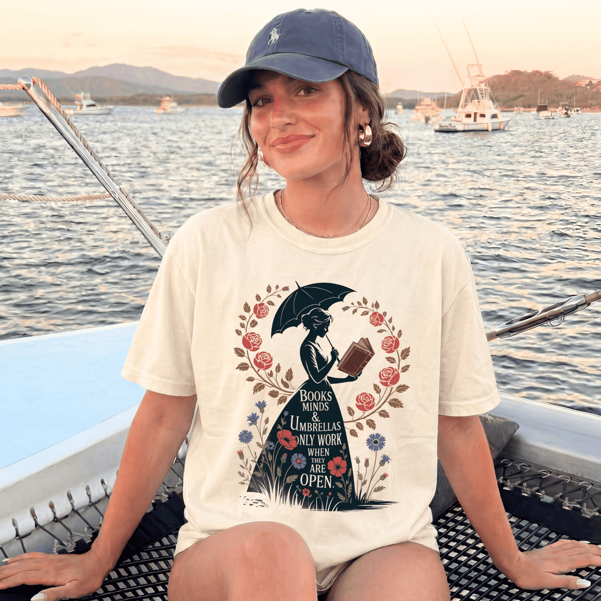 A woman wearing a white t-shirt with a graphic design of a woman reading a book while holding an umbrella, sitting on a boat in a body of water with mountains in the background.