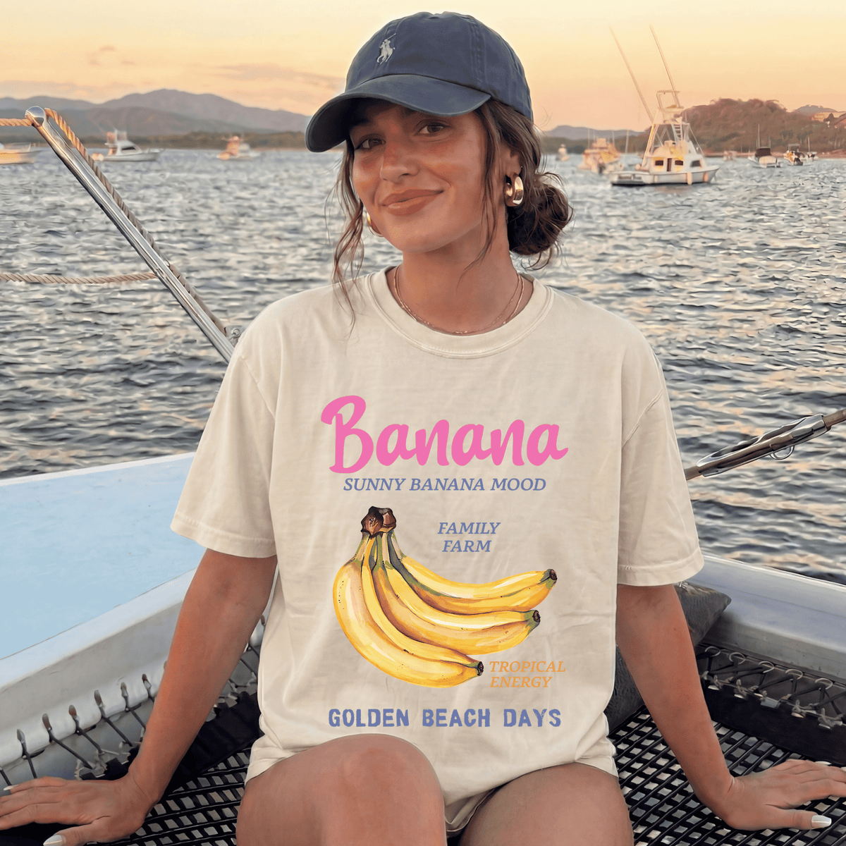 A woman wearing a white t-shirt with a graphic of bananas and text, sitting on a boat in front of a body of water with mountains in the background.