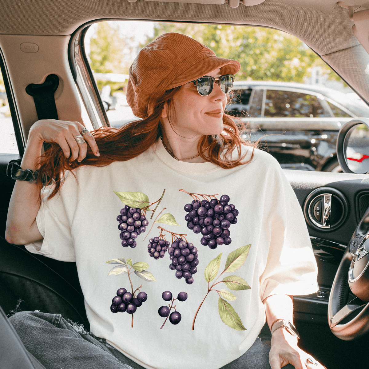 A woman with red hair is sitting in a car, wearing a white t-shirt with a graphic of purple grapes and green leaves.