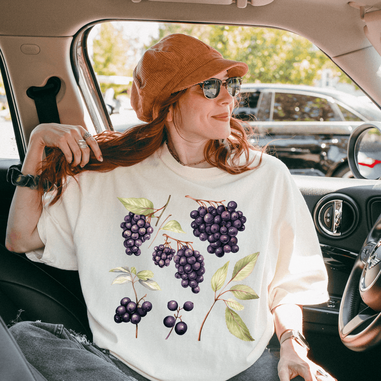 A woman with red hair is sitting in a car, wearing a white t-shirt with a graphic of purple grapes and green leaves.