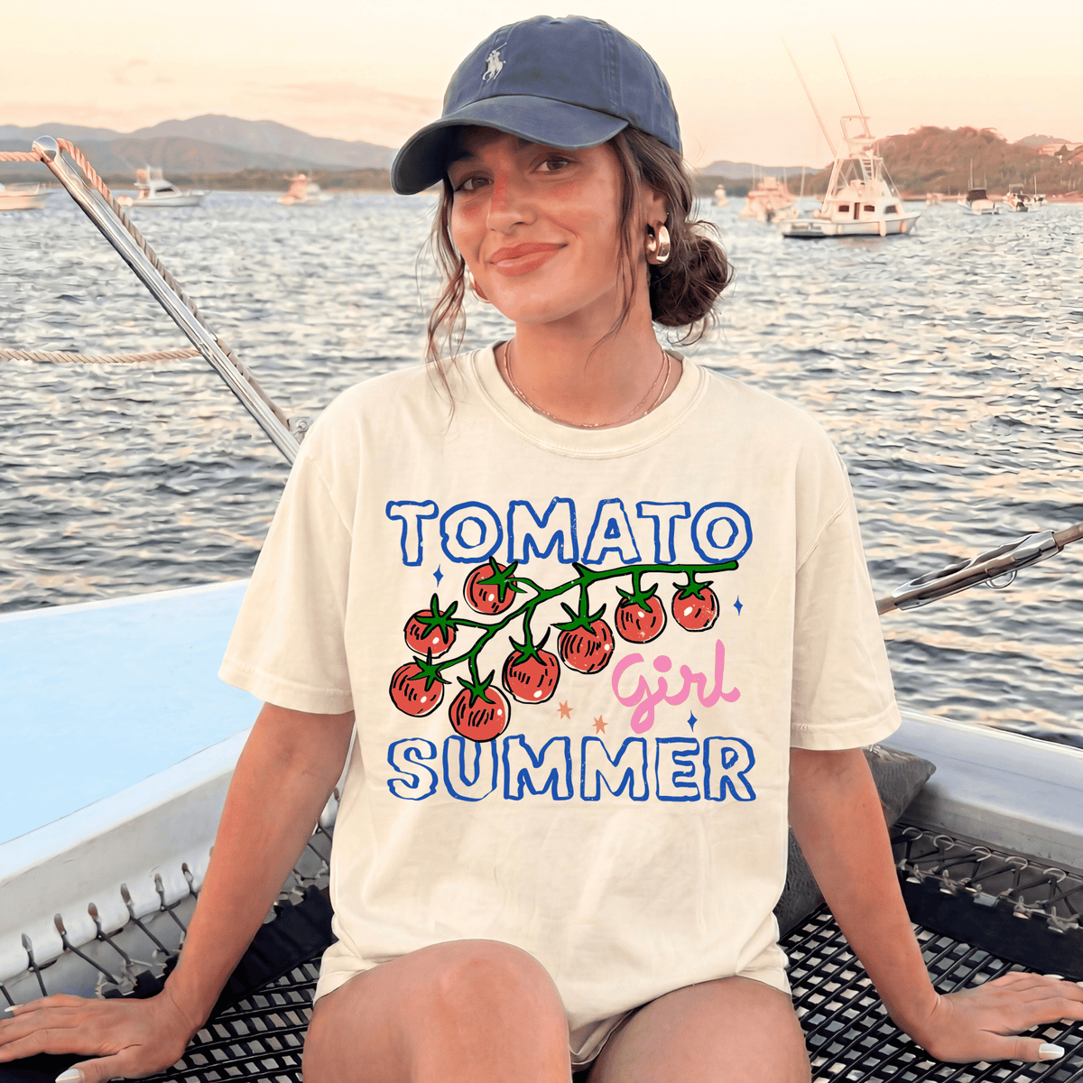 A woman wearing a white t-shirt with a graphic of tomatoes and the text "Tomato girl summer" is sitting on a boat in the water.