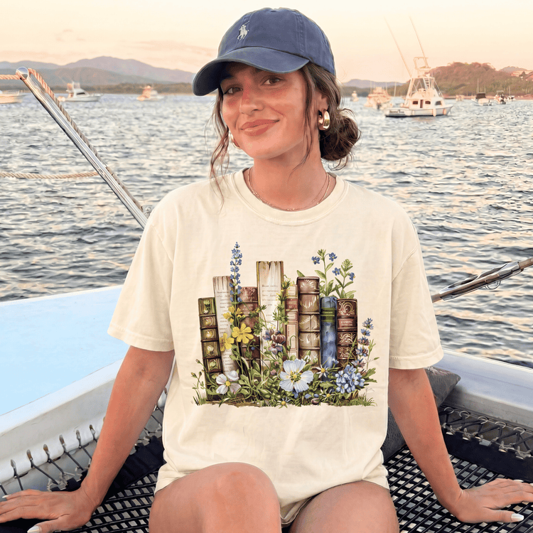 A woman wearing a white t-shirt with a graphic of books and flowers is sitting on a boat in a body of water, with mountains and boats in the background.
