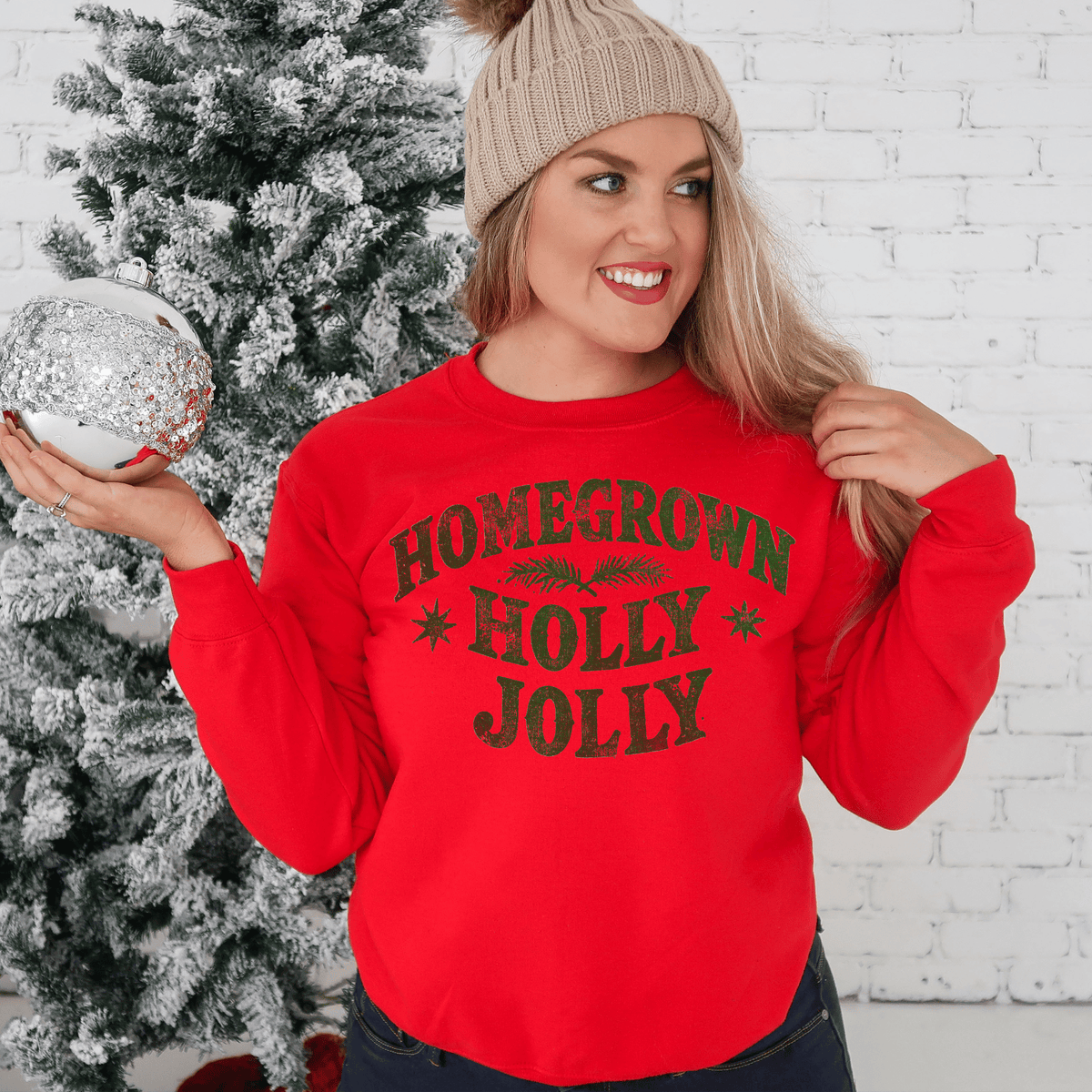 A woman wearing a red sweater with the text "HOMEGROWN HOLY JOLY" stands in front of a Christmas tree, smiling and holding a silver ornament.