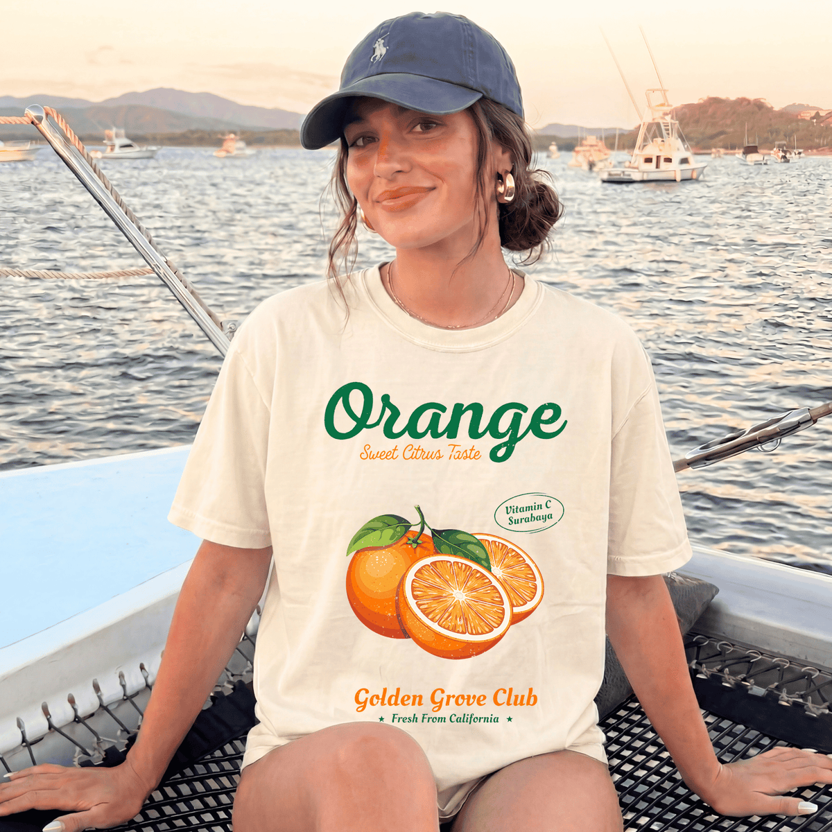 A woman wearing a white t-shirt with an orange graphic and text, sitting on a boat with a scenic background of mountains and other boats.