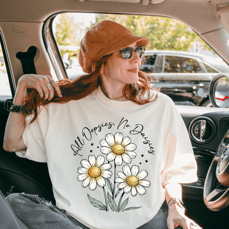 A woman with red hair is sitting in a car, wearing a white t-shirt with a flower design and the text "All Dopsies No Dopsies" printed on it.
