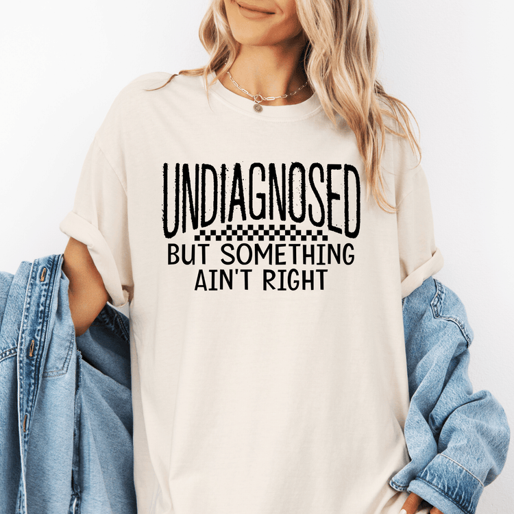 A woman wearing a white t-shirt with the text "UNDIAGNOSED BUT SOMETHING AIN'T RIGHT" printed on it.
