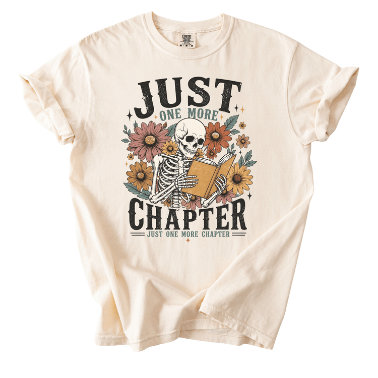 A white t-shirt with a graphic design featuring a skeleton holding a book and surrounded by flowers. The text on the t-shirt reads "JUST ONE MORE CHAPTER" and "JUST ONE MORE CHAPTER".