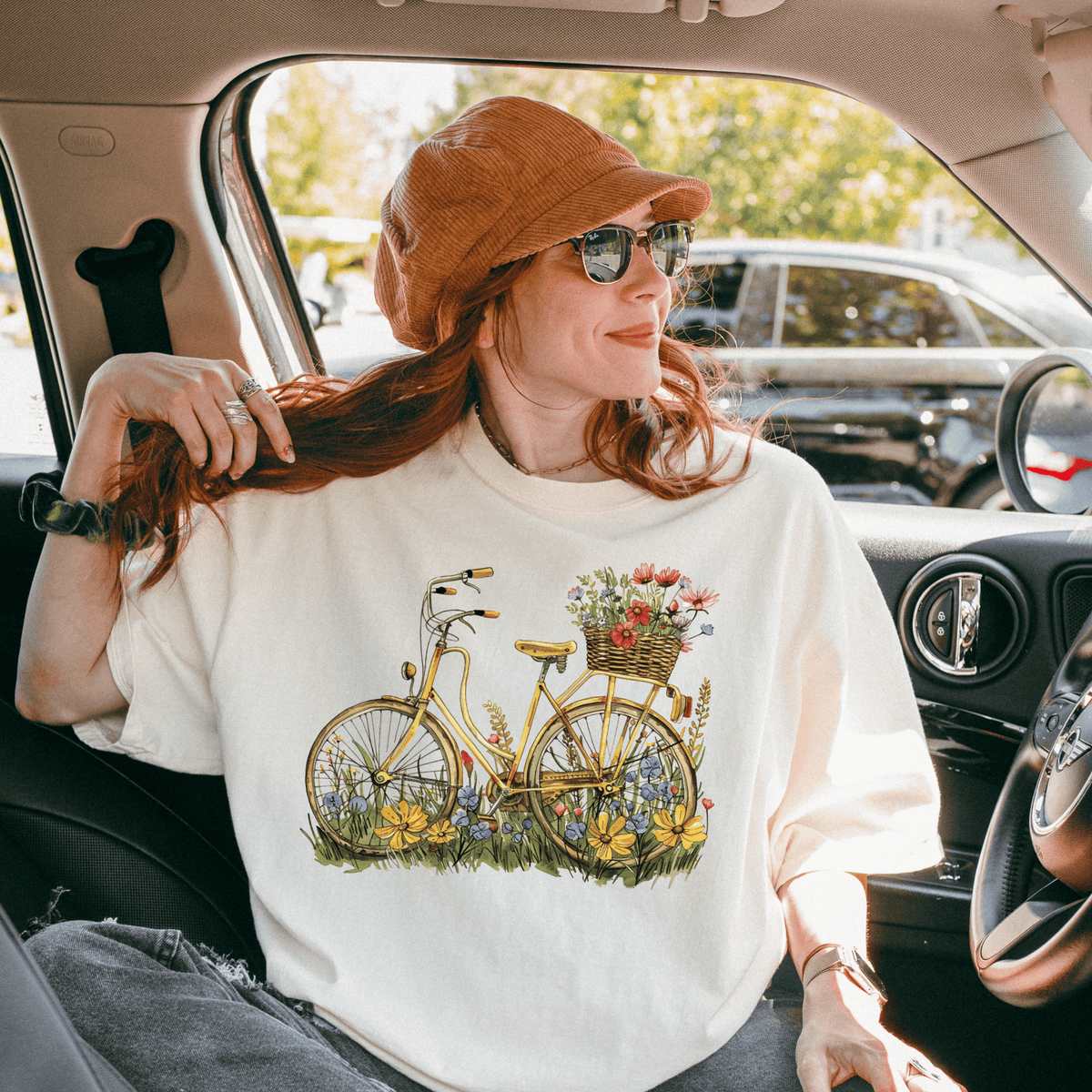 A woman wearing a white t-shirt with a bicycle graphic and flowers is sitting in a car, smiling and looking out the window.