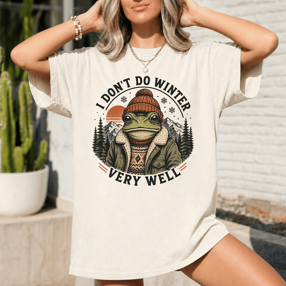 A woman wearing a white t-shirt with a graphic design of a frog wearing a winter hat and jacket, along with the text "I DON'T DO WINTER VERY WELL".