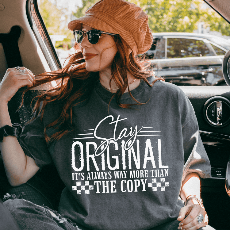 A woman wearing a hat and sunglasses is sitting in a car, looking out the window. She is wearing a gray t-shirt with a text design on it.