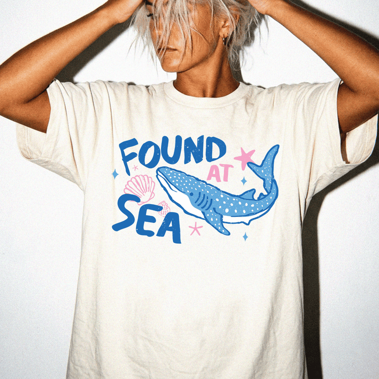A person wearing a white t-shirt with a graphic design of a blue whale and seashells, along with the text "FOUND AT SEA".