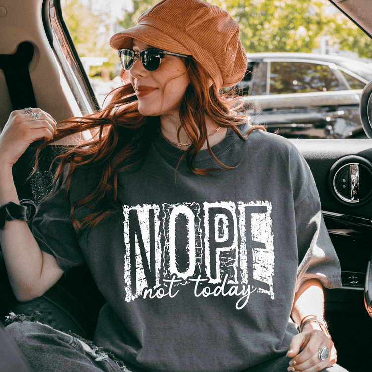 A woman wearing a hat and sunglasses is sitting in the back seat of a car, looking out the window. She is wearing a gray t-shirt with the text "NOPE not today" printed on it.
