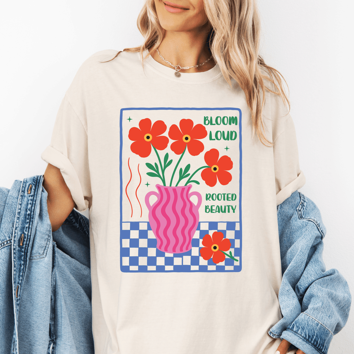 A woman wearing a white t-shirt with a graphic design featuring a pink vase with red flowers and the text "Bloom loud rooted beauty".