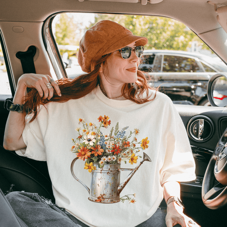A woman with red hair is sitting in a car, wearing a white t-shirt with a flower design and a brown hat.