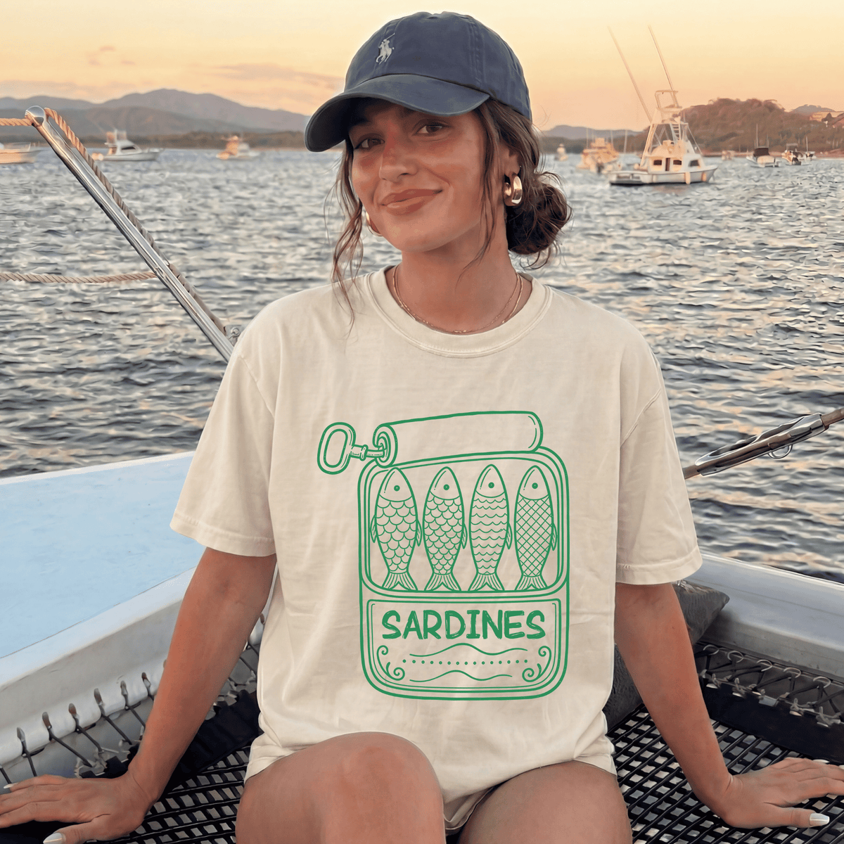 A woman wearing a hat and a t-shirt with a graphic of fish sitting on a boat in a body of water.