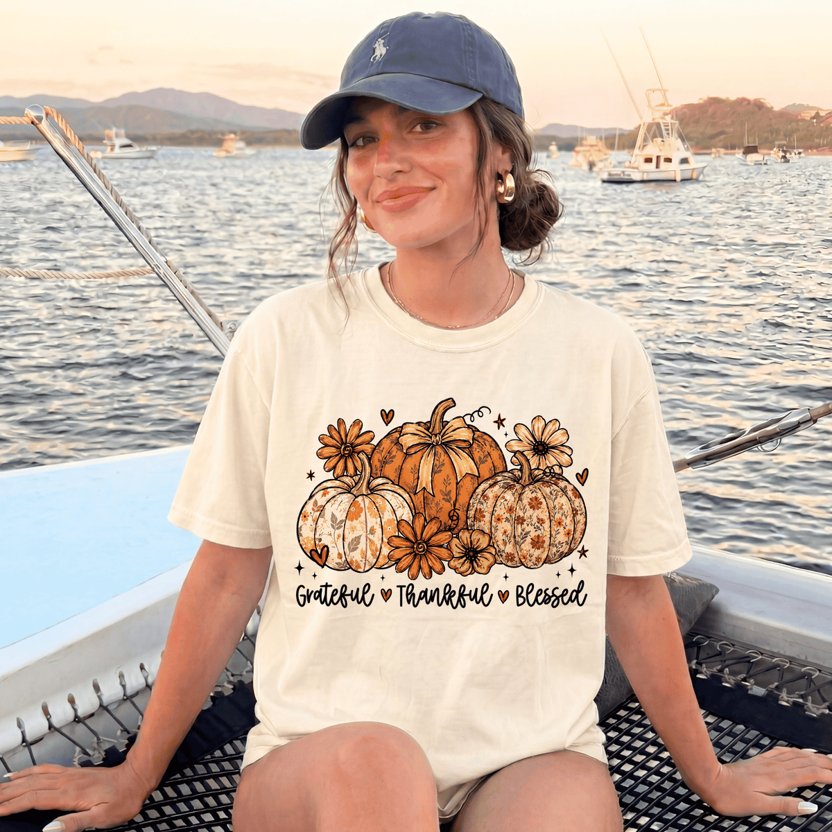 A woman wearing a white t-shirt with a graphic of pumpkins and flowers, sitting on a boat with a scenic background of mountains and boats.