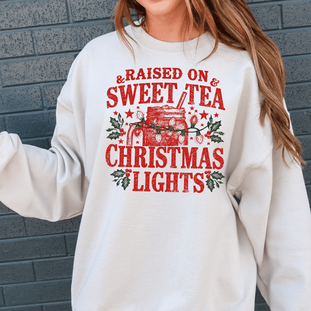 A white sweatshirt with a graphic design featuring a mug with Christmas lights and the text "Raised on & Sweet Tea Christmas Lights" printed on it.