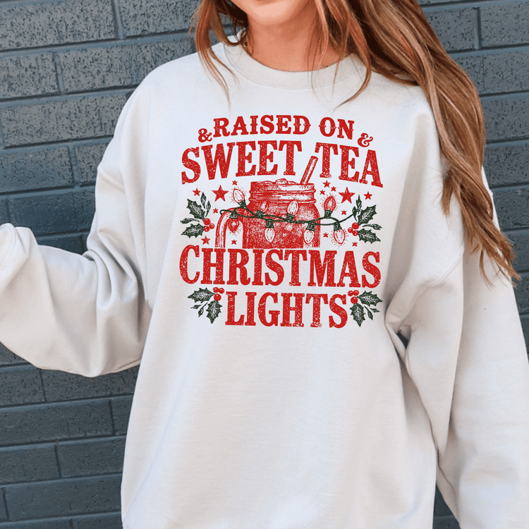 A white sweatshirt with a graphic design featuring a mug with Christmas lights and the text "Raised on & Sweet Tea Christmas Lights" printed on it.