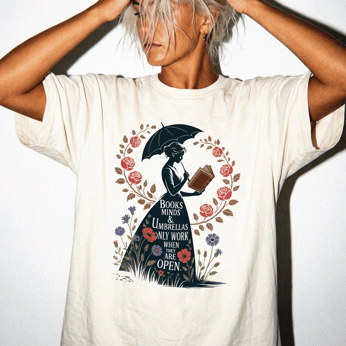 A woman wearing a white t-shirt with a graphic design featuring a silhouette of a woman holding an umbrella and reading a book, surrounded by flowers and leaves.