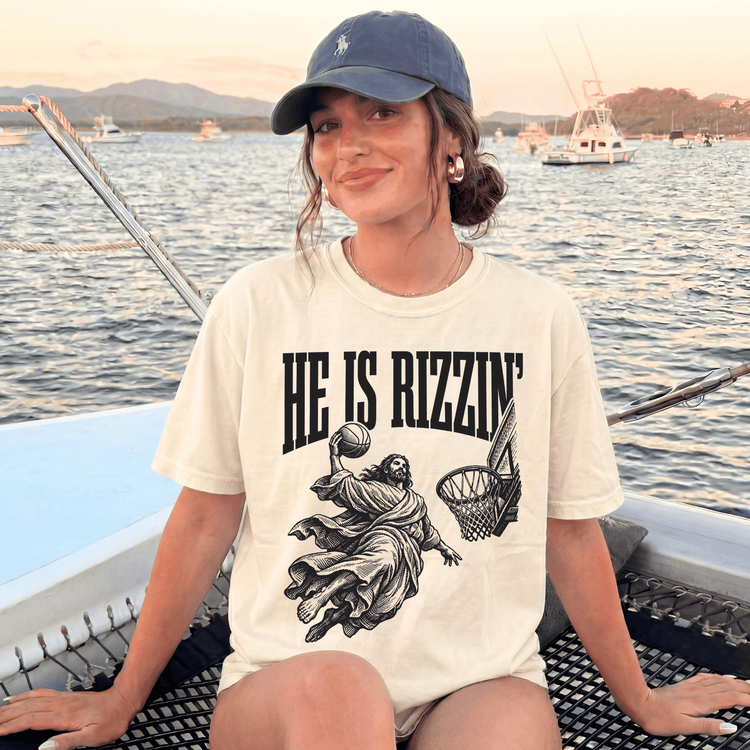 A woman wearing a baseball cap and a t-shirt with a basketball graphic is sitting on a boat in the water, with mountains in the background.