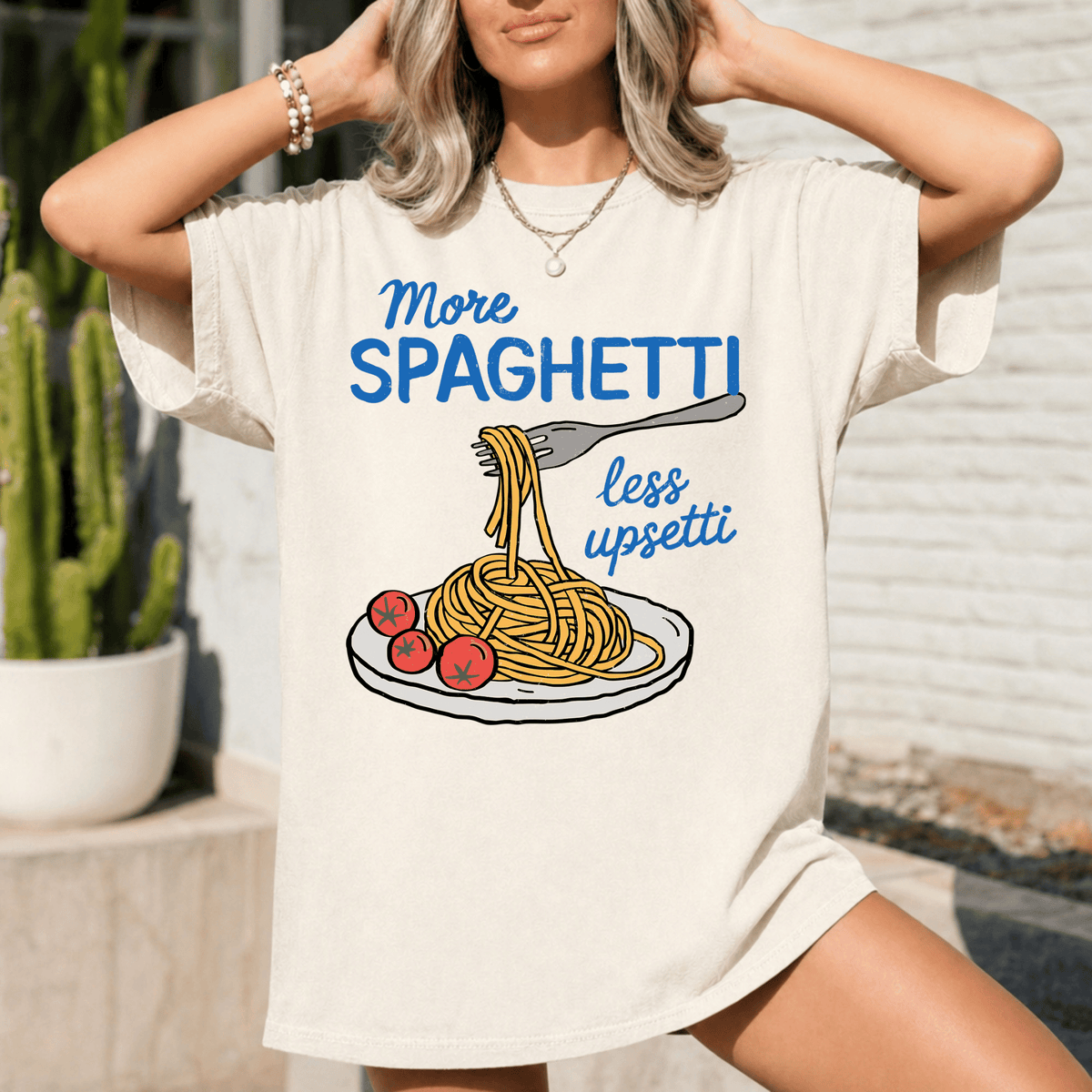 A woman wearing a white t-shirt with a graphic design of spaghetti and tomatoes, along with the text "More spaghetti less upsti".