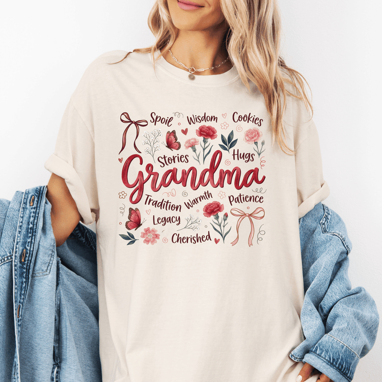A woman wearing a white t-shirt with a graphic design that says "Grandma" surrounded by various words and illustrations such as "Spoils", "Wise", "Cookies", "Hugs", "Stories", "Legacy", "Patience", "Warmth", "Tradition", "Legacy", "Charmed", and "Legacy".