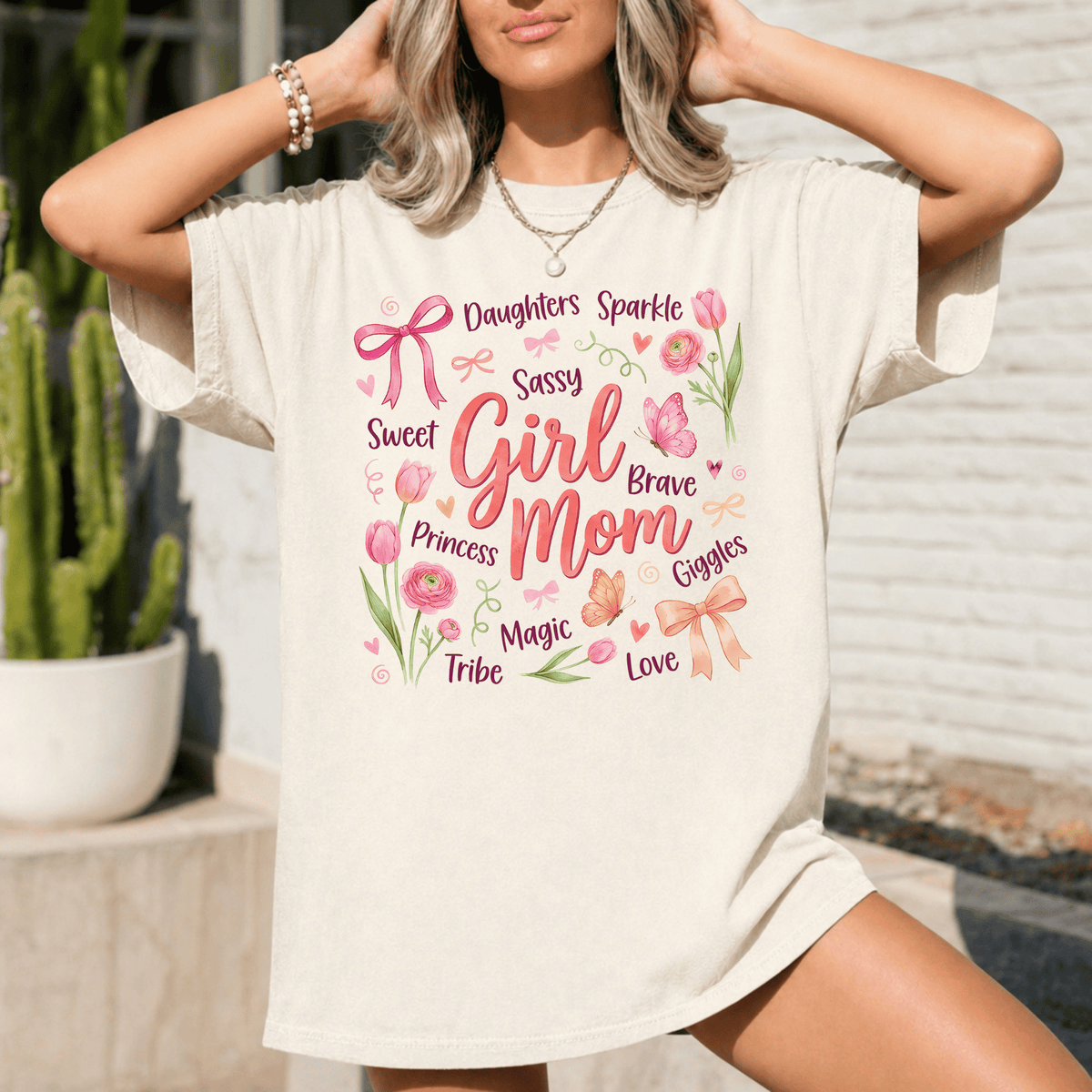 A woman wearing a white t-shirt with a printed design that says "Daughters, Sparkle, Sassy, Sweet, Girl, Brave, Princess, Giggles, Magic, Tribe, Love" surrounded by various flowers and decorative elements.