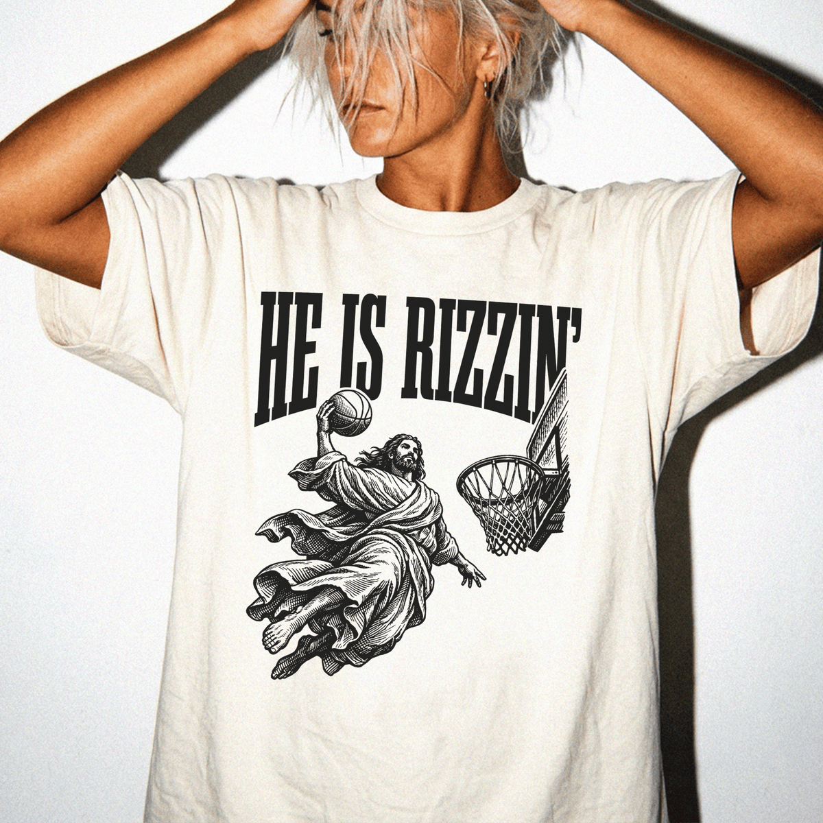 A person wearing a white t-shirt with a graphic design depicting a basketball player in a dramatic pose, with the text "HE IS RIZZIN'".