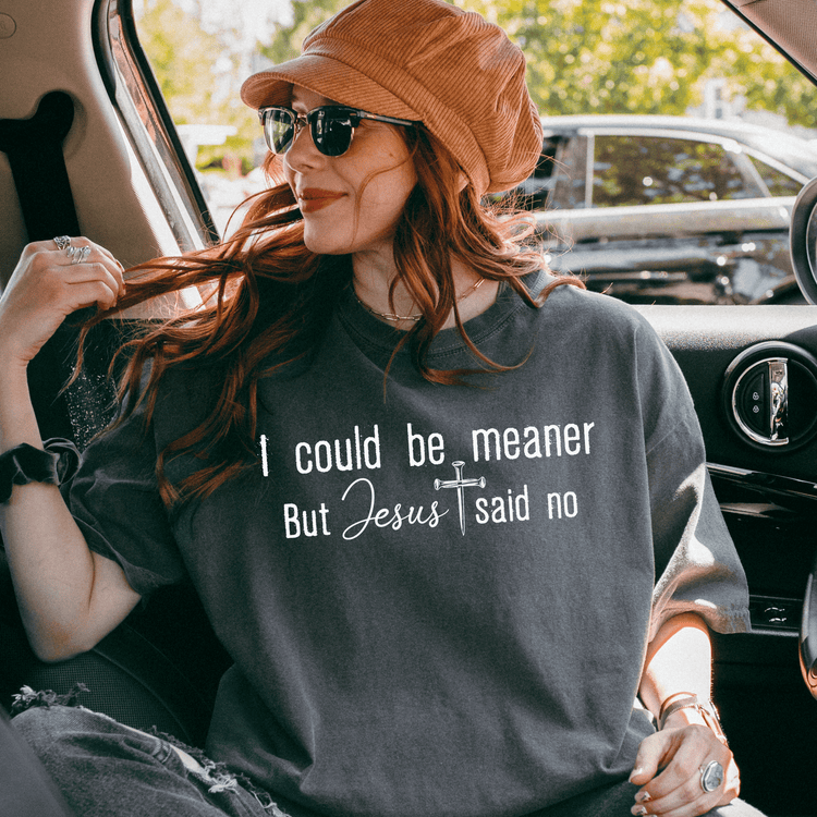 A woman wearing a hat and sunglasses is sitting in the back seat of a car, looking out the window. She is wearing a gray t-shirt with text on it.