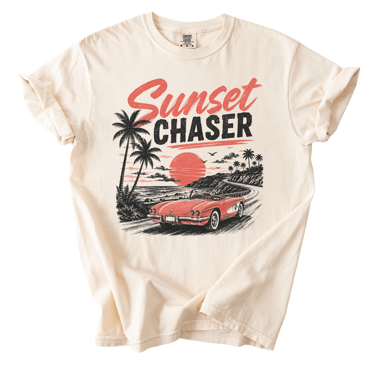 A white t-shirt with a graphic design depicting a vintage car on a road with palm trees and a sunset in the background.