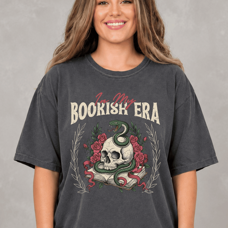 A woman wearing a gray t-shirt with a graphic design featuring a skull, a snake, and a book.