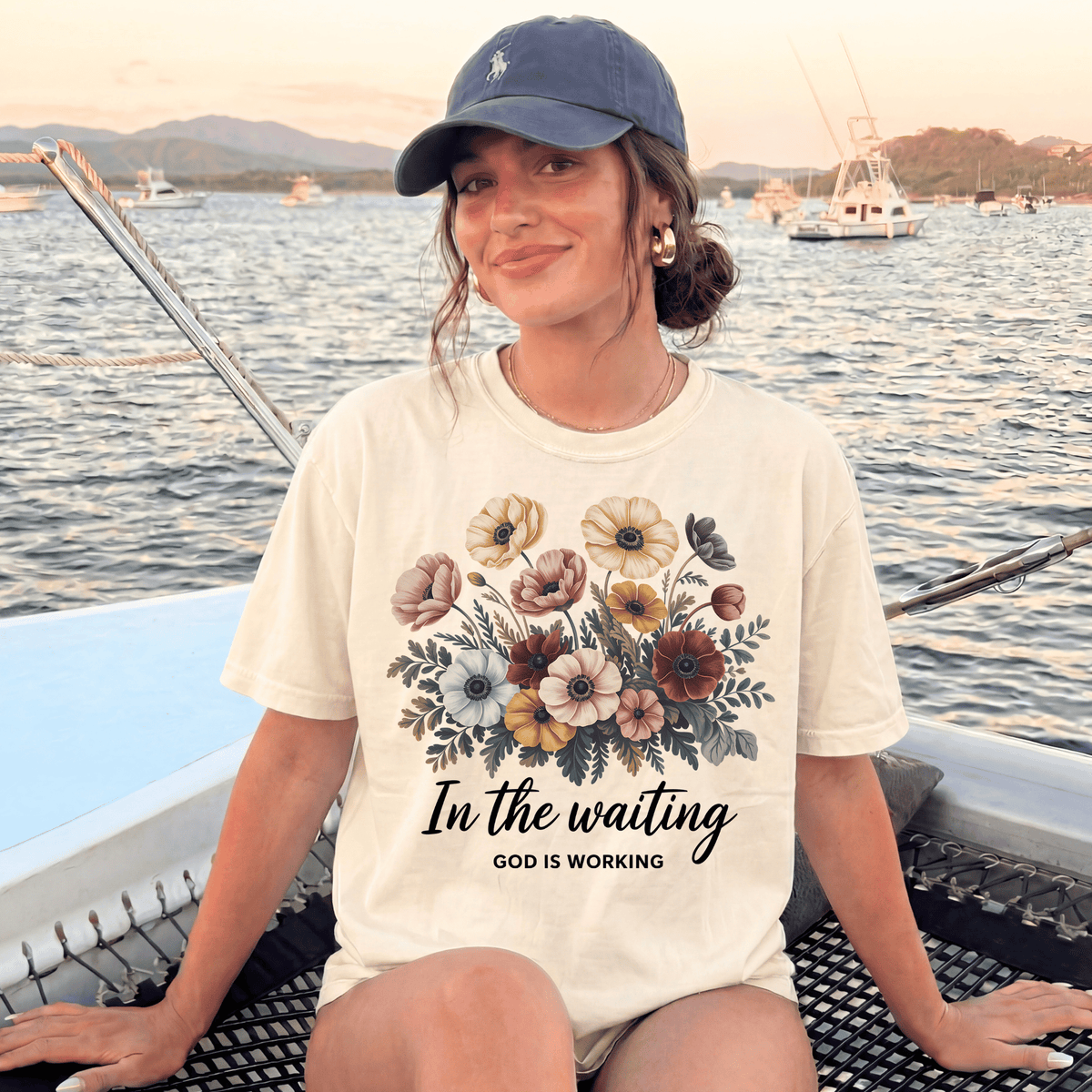 A woman wearing a white t-shirt with a floral design and the text "In the waiting God is working" is sitting on a boat in the water.
